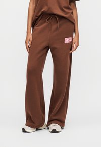 Καφέ φόρμα από μαλακό ύφασμα με άνετη εφαρμογή. Διαθέτει ελαστική μέση με κορδόνι και ροζ λογότυπο Nike στην αριστερή πλευρά.