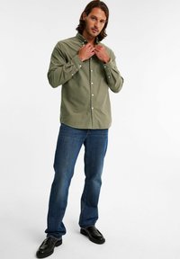 Olivgrön långärmad skjorta med knappar och ficka, i kombination med blå jeans och svarta skor, står framför en enkel bakgrund.