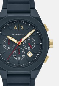 Muška kronografska sat s tamnoplavim silikonskim remenom i okruglim mat kućištem. Ima zlatne i crvene akcentu, tri subdijela i prikaz datuma.
