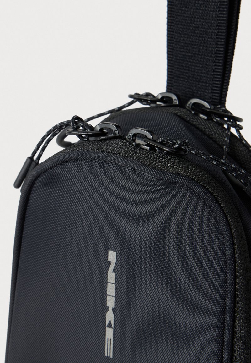 Nærbillede af en sort stofslyngetaske med dobbelte lynlåse og en rem, der viser mærket Nike med lodrette sølvbogstaver.