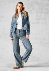 Veste en denim bleu clair au design ajusté, associée à un pantalon large assorti. Chemise blanche en dessous, complétée par des baskets beige.