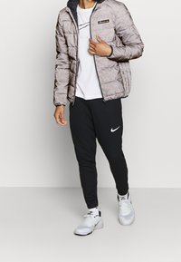 Mand iført en lys grå mønstret vinterjakke, hvid T-shirt, sorte Nike sweatpants, hvide Nike sneakers, stående mod en ensfarvet baggrund.