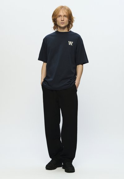 Jeune homme aux cheveux roux mi-longs porte un t-shirt bleu marine ample avec un logo blanc, un pantalon noir et des chaussures noires, debout les mains dans les poches.