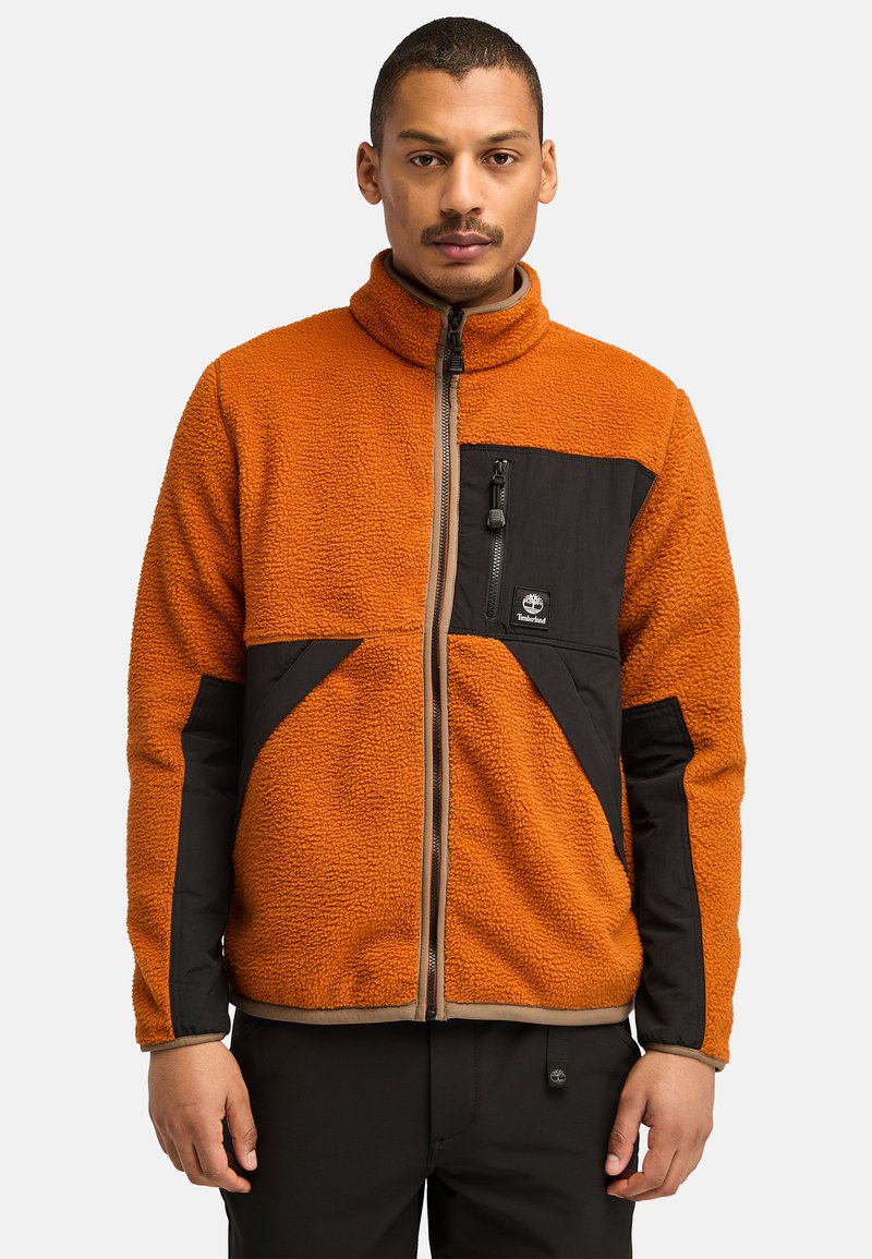Fleecejacke mit orangem Körper, schwarzen Stoffakzenten, einem hohen Kragen und einer mit Reißverschluss versehenen Brusttasche. Hat eine strukturierte Außenseite.