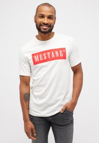 Balta medvilninė marškinėliai su dideliu raudonu stačiakampiu "MUSTANG" logotipu baltu tekstu; apvalia iškirpte, trumpomis rankovėmis, švelnios tekstūros.