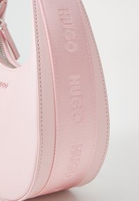Sac à épaule rose en cuir lisse, avec une forme arrondie, un logo en relief et une sangle texturée avec marquage.