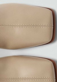 Beige lædersko med firkantet tå, der har en glat overflade og synlige sømme langs midten. Brune detaljer ved sålen.