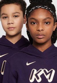 Sudaderas con capucha moradas con el logo "KM" y la marca Nike. Tejido suave, puños acanalados. Dos niños con trenzas y rizos cortos miran hacia adelante.
