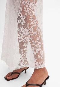 Piernas con falda de encaje floral blanco transparente y sandalias de tacón negro con tiras sobre un fondo blanco.