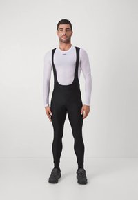 Salopette da ciclismo nera con bretelle regolabili, realizzata in materiale elastico, abbinata a una maglietta a maniche lunghe bianca aderente con un logo.