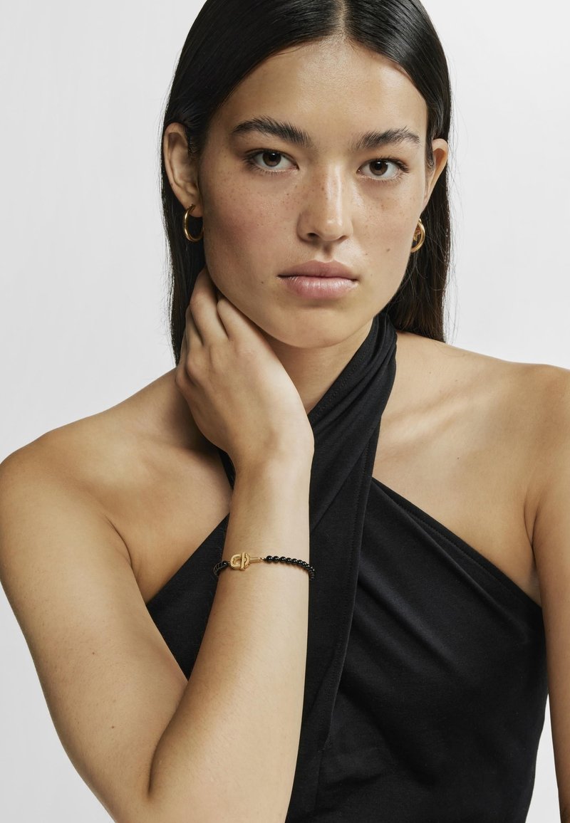 Top negro sin mangas con un diseño retorcido, combinado con una delicada pulsera de cuentas negras con un cierre dorado. Piel suave con pecas naturales.