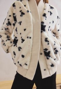 Faux fur jacket in cream with black speckles, featuring a shawl collar and ribbed cuffs. Button closure visible at the bottom.