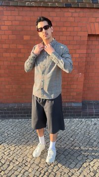 Homme portant des lunettes de soleil, une chemise à carreaux, un short noir, des chaussettes blanches et des baskets, ajustant son col tout en se tenant dans une rue pavée près d'un mur en briques.