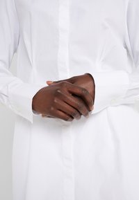 Chemise blanche à manches longues avec col classique, caractérisée par une texture lisse et des poignets avec boutons. Les mains sont croisées devant.