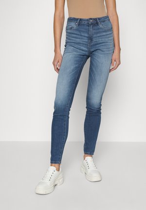 Jeans de ganga azuis de cintura alta, com design ajustado, cor desgastada, estilo de cinco bolsos, combinados com ténis brancos. Textura suave.