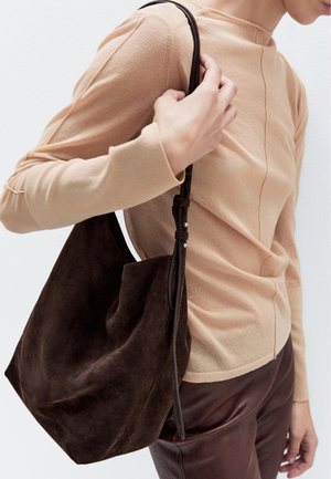 Borsa a spalla in suede marrone con un design morbido e sformato, tenuta da una mano. Sono visibili un top in maglia beige chiaro e pantaloni in pelle scura.