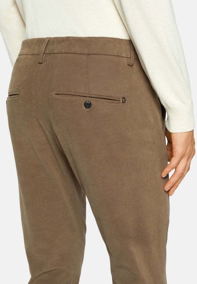 Pantalons marron en tissu texturé, avec deux poches arrière à boutons et une coupe ajustée. Un pull couleur ivoire est partiellement visible.