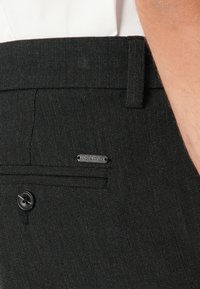 Gros plan sur la poche d'un pantalon noir texturé, avec un bouton et une étiquette en métal de la marque Jack & Jones, porté avec une chemise blanche.