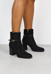 Botines de ante negros con punta cuadrada, tacón bajo y acento de correa de cuero negro. Presenta una cremallera lateral para el cierre.