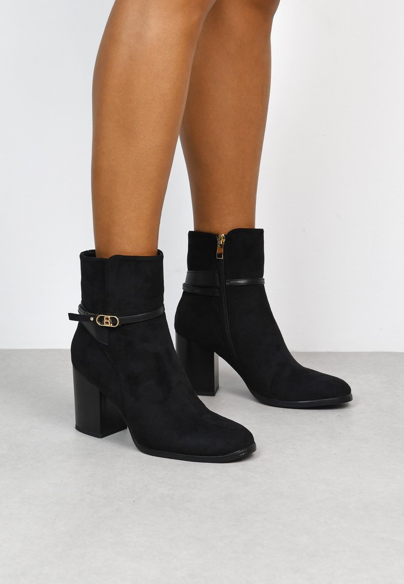 Botines de ante negros con punta cuadrada, tacón bajo y acento de correa de cuero negro. Presenta una cremallera lateral para el cierre.