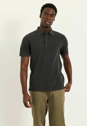 Junger Mann trägt ein dunkles Poloshirt und olivgrüne Hose, steht vor einem einfarbigen hellen Hintergrund und schaut in die Kamera.