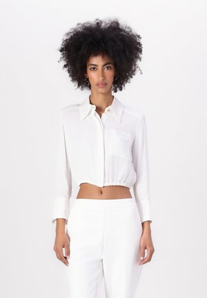 Camicia bianca a maniche lunghe, con colletto a punta e bottoni, in tessuto leggero e orlo inferiore arricciato. Piccola tasca sul petto.