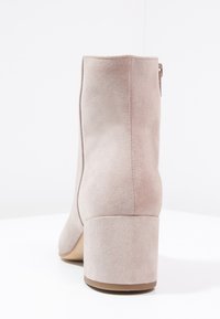 Botte de cheville en suède beige avec un talon bloc et une fermeture éclair à l'arrière, vue de derrière sur un fond blanc.