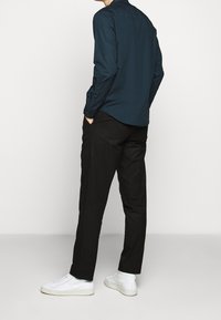 Mężczyzna w ciemnoniebieskiej koszuli z długim rękawem, czarnych spodniach i białych sneakersach, stojący z rękami w kieszeniach na jednolitym jasnym tle.