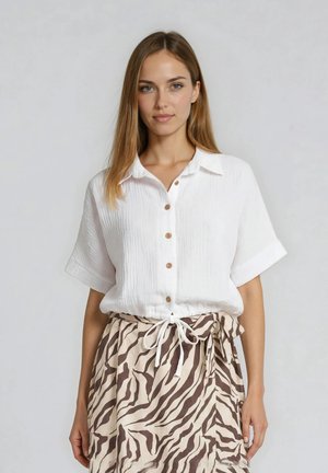 Femme portant une chemise blanche à manches courtes boutonnée et une jupe beige avec des rayures zébrées marron, nouée à la taille avec un cordon blanc.