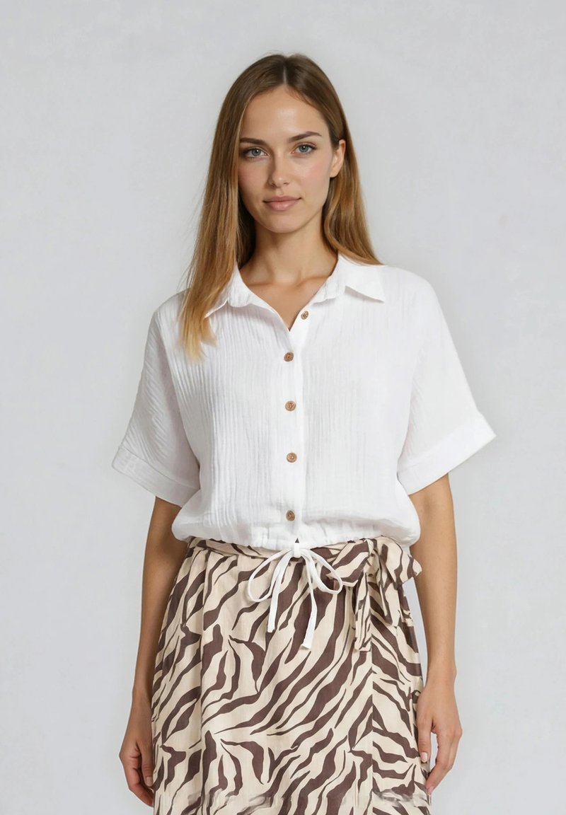 Femme portant une chemise blanche à manches courtes boutonnée et une jupe beige avec des rayures zébrées marron, nouée à la taille avec un cordon blanc.