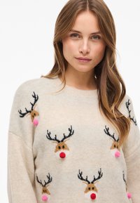 Pull crème avec motifs de rennes aux têtes brunes, bois noirs et pompons roses. Texture douce, col rond et coupe ample.