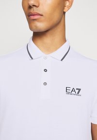 Bílá polo košile s šedými akcenty, dvouknoflíková léga a logo EA7 na hrudi. Vyrobena z texturovaného materiálu, s klasickým límečkem.