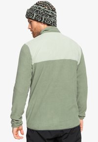 Zielona polarowa kurtka z jasnzieloną górną częścią i teksturowanym wykończeniem. W połączeniu z czapką dzianinową w czarno-zielone wzory.