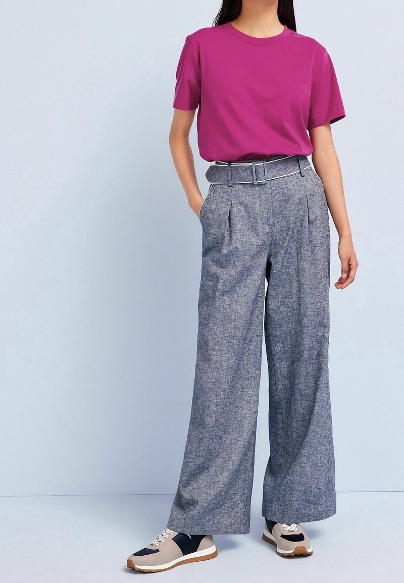 Personne portant une chemise rose vif à manches courtes, un pantalon gris taille haute à jambes larges avec ceinture, ainsi que des baskets beige, noires et blanches.