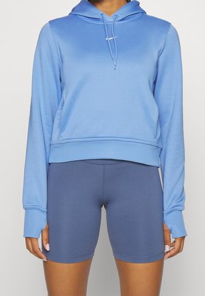 Personne portant un sweat à capuche bleu clair avec cordons et un cuissard de vélo ajusté bleu foncé, debout devant un fond uni.