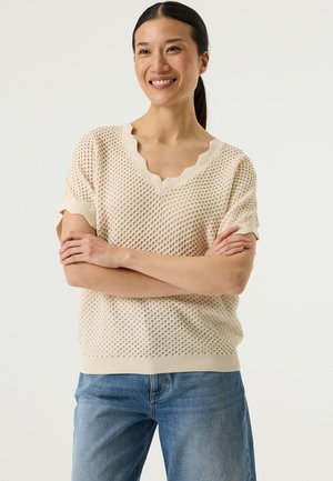 Pull beige en maille avec un design texturé à tissage ouvert, encolure en V festonnée et manches courtes, associé à un jean en denim bleu clair.