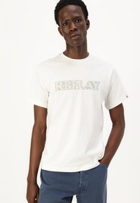 Hvit bomull T-skjorte med et grått "REPLAY"-logo i en fet skrifttype. Rund hals og korte ermer, sammen med marineblå bukser.