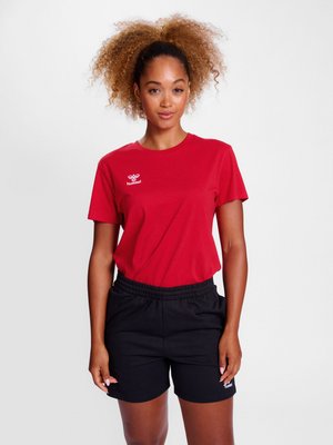 Młoda kobieta z kręconymi włosami, ubrana w czerwony t-shirt marki hummel oraz czarne szorty z elastyczną talią, stoi na tle jednolitego białego tła.