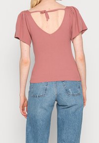 Geribbelde, rozenkleurige top met pofmouwen en een diepe V-rug, met een strikdetail. Gecombineerd met blauwe denim wijde pijpen jeans.