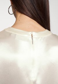 Top in satin avorio chiaro con scollatura rotonda, dotato di una discreta zip sul retro e dalla texture liscia. Design semplice con dettagli minimali.