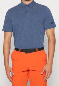 Laivastonsininen lyhythihainen pikeepaita, jossa on kaulus, kaksi nappia ja vasemmassa hihassa kirjailtu logo, yhdistettynä oransseihin housuihin ja mustaan vyöhön.