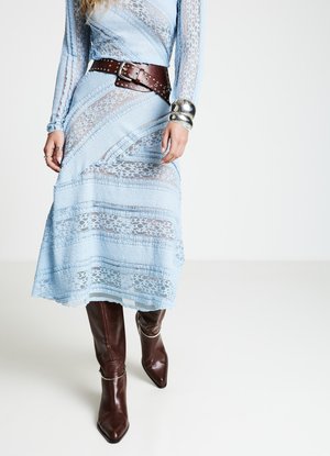 Hellblaues Spitzenkleid mit horizontalen Mustern, geschnürtem braunen Ledergürtel und silbernem Akzentarmband. Braune kniehohe Stiefel mit spitz zulaufender Schuhspitze.