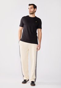 Sort t-shirt med korte ærmer og gult logo, parret med cremefarvede bukser med sorte side-striber, og mørke sneakers med en tekstureret overflade.