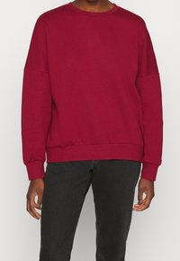Sudadera burdeos con cuello redondo, hombros caídos, mangas largas y ribete en el dobladillo. Textura suave, diseño casual y corte holgado.