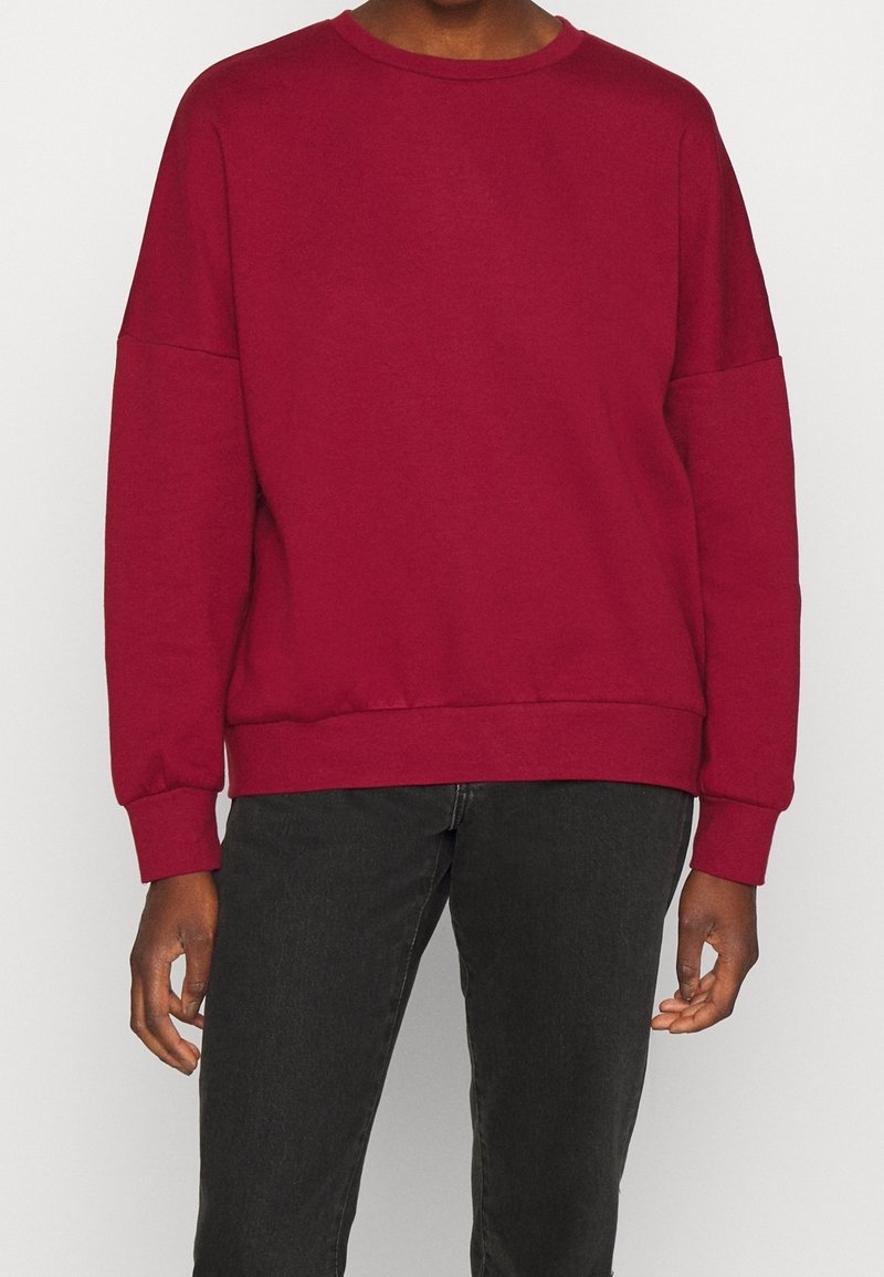 Sudadera burdeos con cuello redondo, hombros caídos, mangas largas y ribete en el dobladillo. Textura suave, diseño casual y corte holgado.
