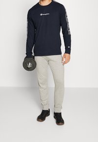 Námořnické tričko s dlouhým rukávem a bílým logem, šedé tepláky a černé sportovní boty, držící černou činku. Jednoduchý, neformální vzhled pro aktivní odívání.