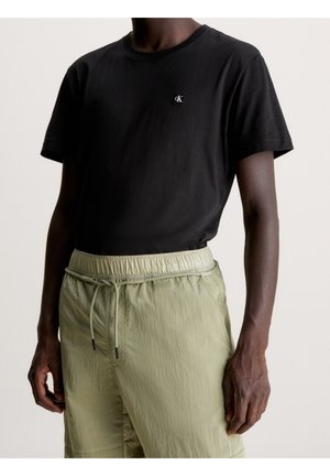 Homme portant un t-shirt noir à manches courtes avec un petit logo "cK" et un pantalon vert clair à cordon de serrage, cadré à la poitrine et à mi-cuisse.