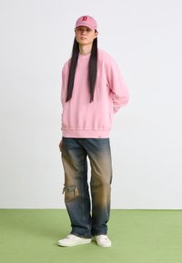 Ροζ φλις φούτερ, σκισμένα τζιν με σπασμένες άκρες, λευκά αθλητικά παπούτσια και ροζ καπέλο με κόκκινο λογότυπο. Στεκόμενος σε πράσινο πάτωμα.