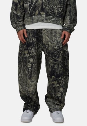 Persona che indossa pantaloni larghi con motivo mimetico e giacca abbinata con T-shirt bianca e scarpe da ginnastica bianche su uno sfondo semplice.