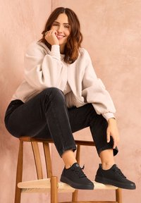 Zapatillas de goma negras con puntas redondeadas y diseño de cordones; combinadas con jeans oscuros y una chaqueta corta de mangas abullonadas en color crema.